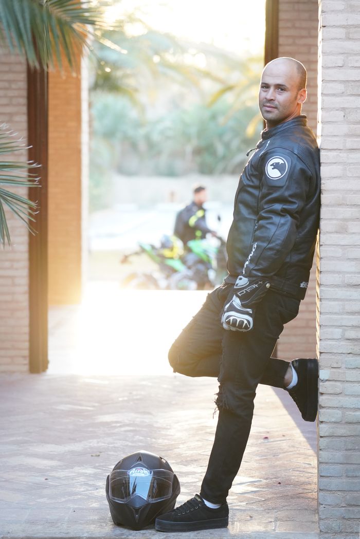 Biker in leather jacket stands against wall outdoors with helmet and gloves, exuding style and confidence.
