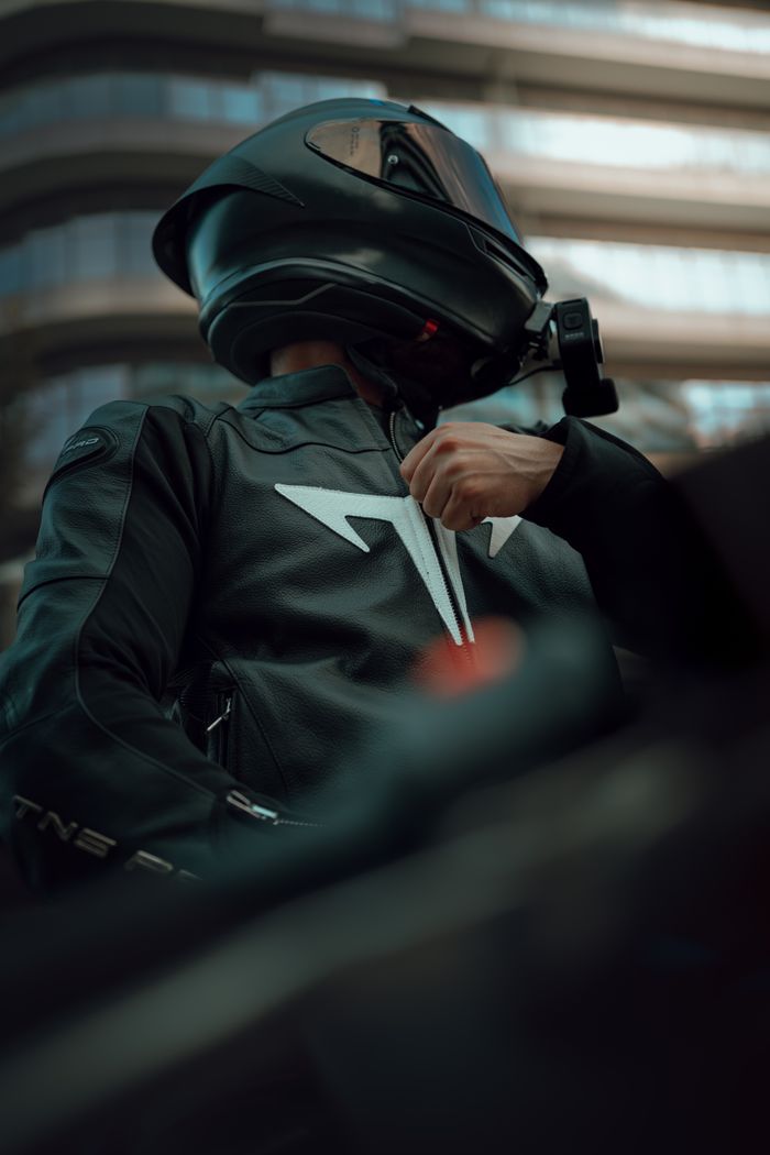 Motorcyclist in black attire adjusting jacket, low angle view in urban setting.