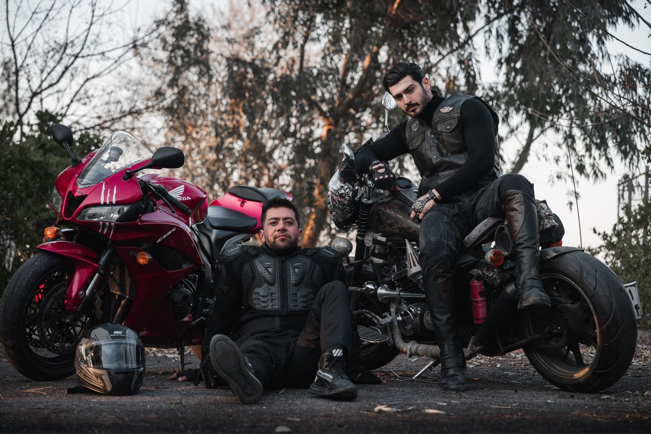 Two bikers in gear pose confidently with motorcycles on an outdoor setting.