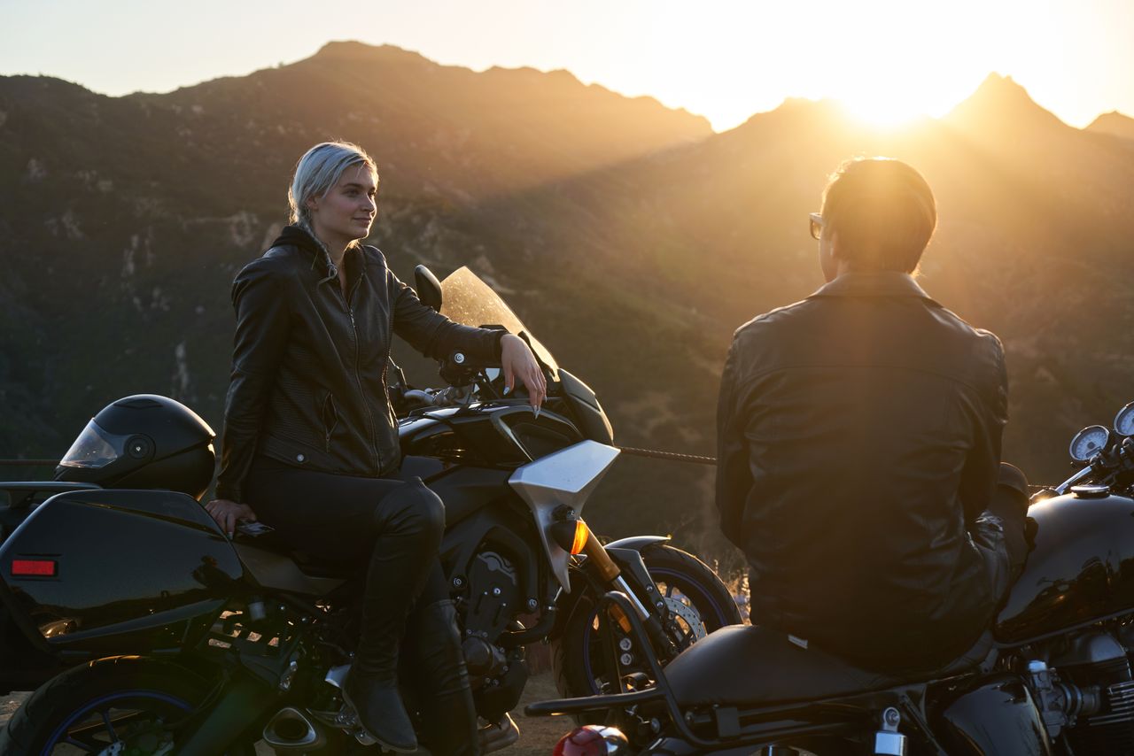 A biker couple wearing leather jackets, sitting on motorcycles at sunset with mountains in the background.